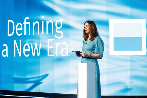 Synnøve Seglem på scenen under årskonferansen 2024, tittelen defining a new era er godt synlig i bakgrunn. Synnøve har på en turkis kjole