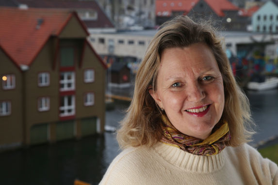 Ingvild Sæther i Altera med hvit strikkegenser og tørkle i halsen. I bakgrunnen havnen og et gult hus med røde karmer.