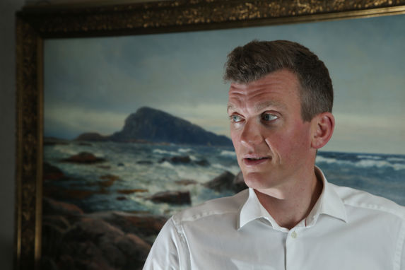 Knut Nikolai Tønnevold Ugland i hvit skjorte, foran et maleri av hav og holmer