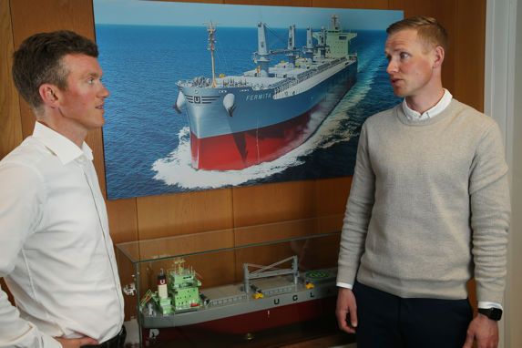 Knut Nikolai Tønnevold Ugland og en mannlig kollega foran en skipsmodell og et foto av et rødt og hvitt skip. 