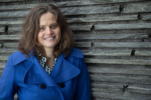 alt="Simone Møkster i koboltblå trenchcoat foran en murvegg "
